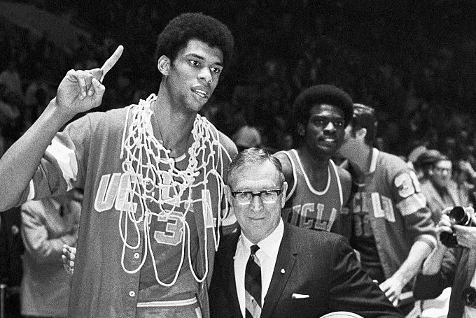 a black and white photo of lew alcindor, wearing a basketball jersey and a basketball net around his neck while holding up a single finger, and john wooden, wearing a suit, glasses, and holding a basketball