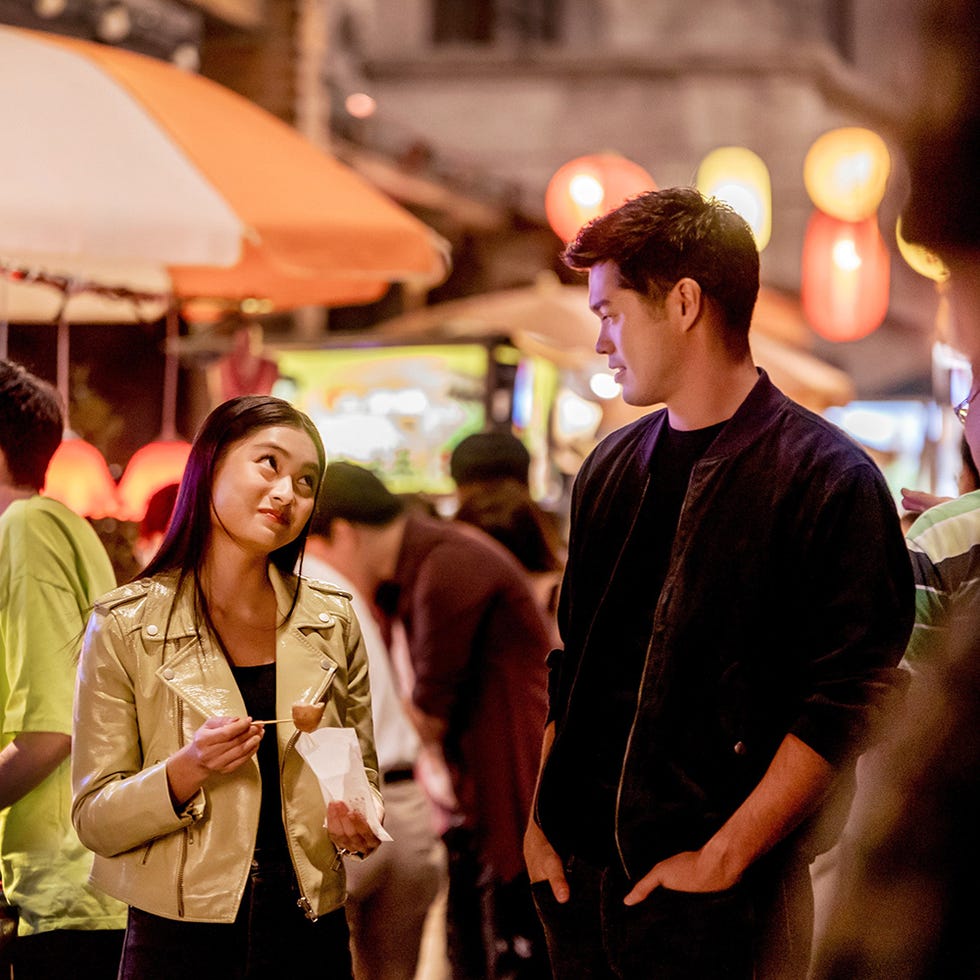 in a vibrant night market, a woman in a shiny gold jacket holds a food item on a stick while engaging in conversation with a man standing beside her in a vibrant night market, a woman in a shiny gold jacket holds a food item on a stick while engaging in conversation with a man standing beside her