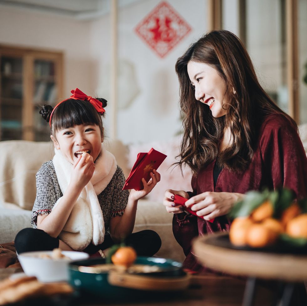 lovely daughter enjoying traditional snacks while helping her mother to prepare red envelops lai see at home for chinese new year