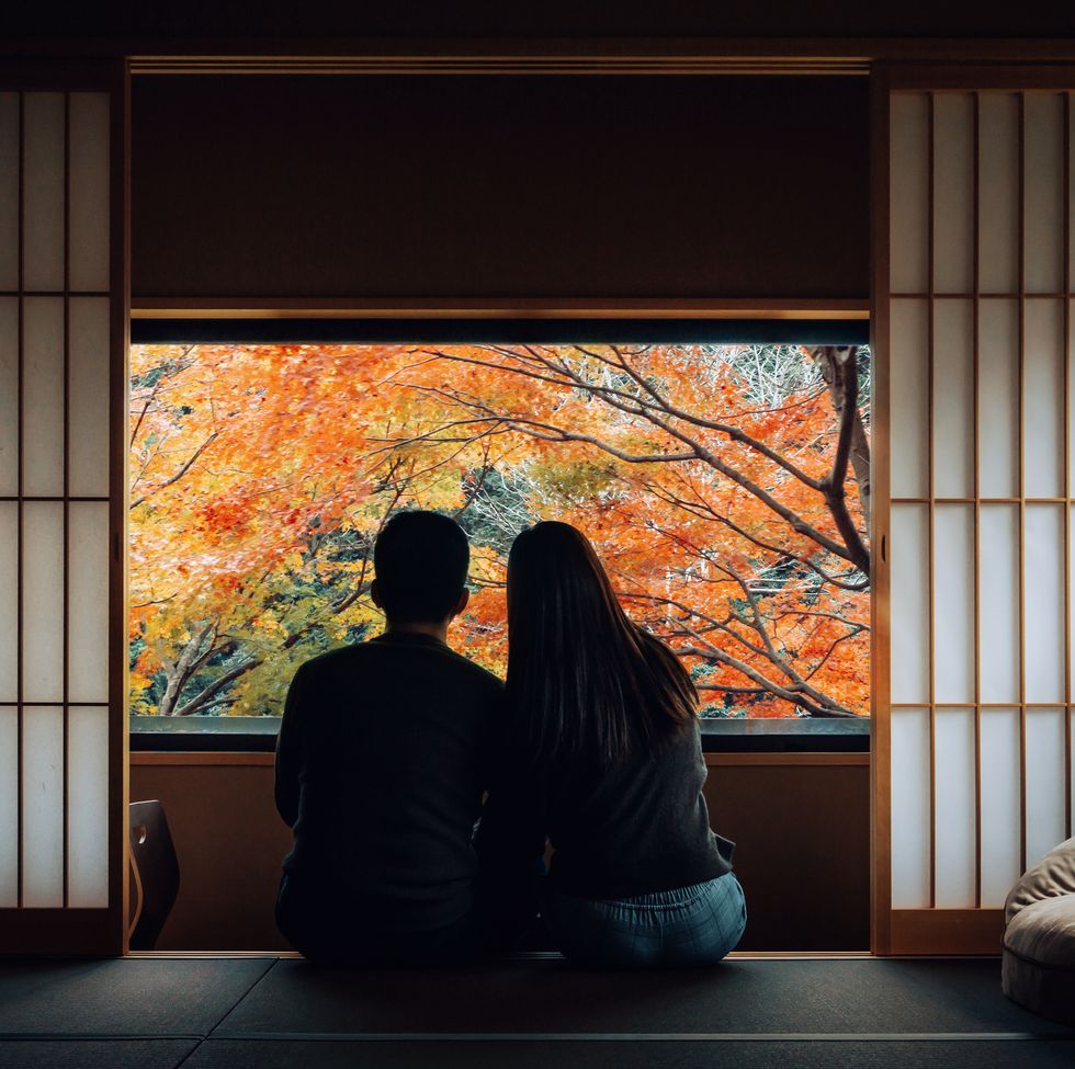 a loving young asian couple sitting side by side by the window in a traditional japanese style apartment, chatting and enjoying the beautiful nature autumn scenics during the day