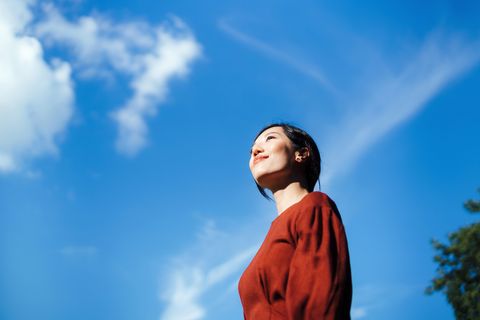 low angle portrait of beautiful smiling young asian woman standing against beautiful clear blue sky, looking up to sky enjoying nature and sunlight freedom in nature connection with nature