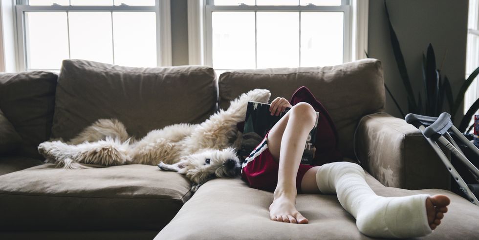 low section of boy with fractured leg reading book while lying by dog on sofa at home
