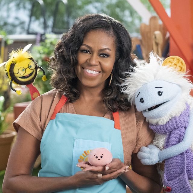 michelle obama, wearing a blue apron, stands in a rooftop garden posing for the camera and holding a pink, round mochi puppet a bee puppet wearing a red tie hovers over her shoulder while a furry white and blue puppet with frozen waffle ears embraces her side