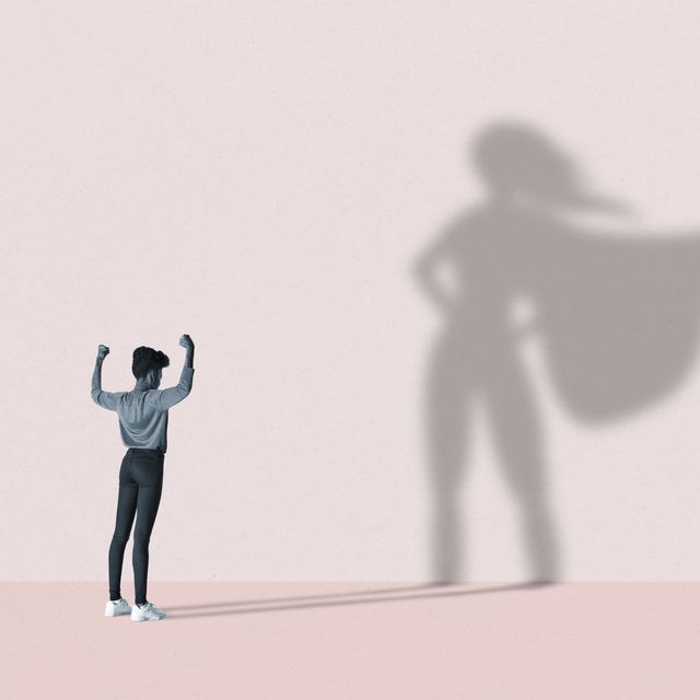 young woman flexing muscles in front of large superhero shadow on pink background