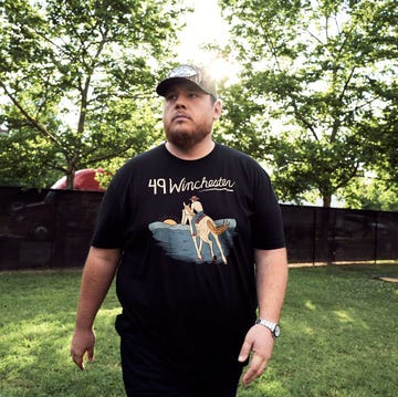 luke combs walks in a grass field, he looks to the left and wears a black graphic t shirt and pants with a baseball cap, sunlight filters through trees behind him