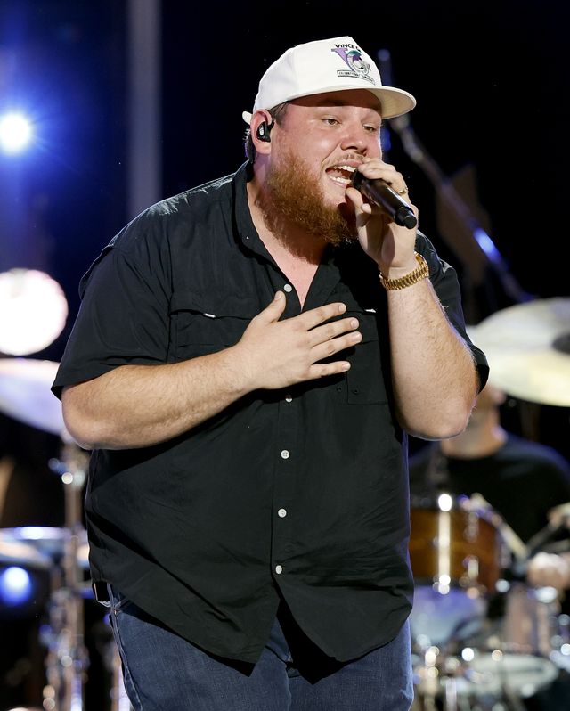 luke combs on a stage singing into a microphone, standing in front of a set of drums, wearing a black shit, jeans, and a white baseball cap
