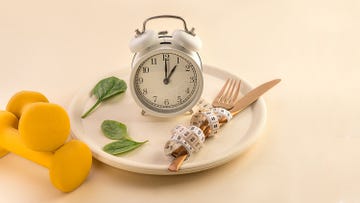 healthy food composition with cutlery, plate, measuring tape, yellow dumbbell, green vegetable salad leaves and alarm clock on pastel beige background dieting and keep fit close up copy space front view