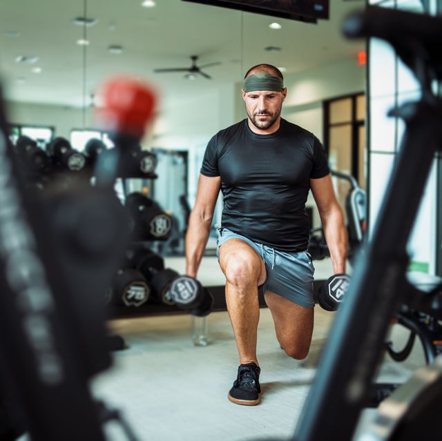 a fit and healthy middle aged caucasian man does a lunge while holding two weighted dumbbells he is concentrating on working out in an indoor gym