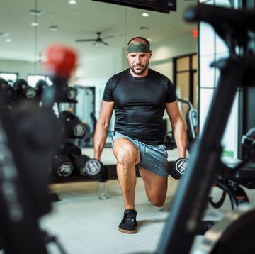 a fit and healthy middle aged caucasian man does a lunge while holding two weighted dumbbells he is concentrating on working out in an indoor gym