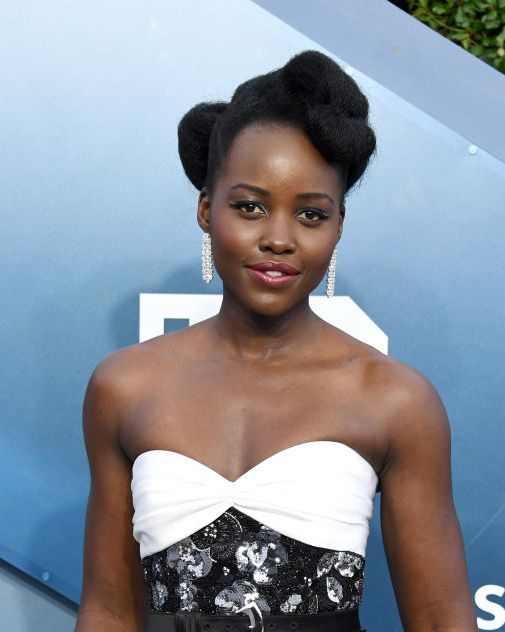 lupia nyong'o stands on the red carpet, wearing a strapless dress with a white top and black and white floral patterned waist and skirt, with her hair up she wears long sparkly earrings and pink lipstick