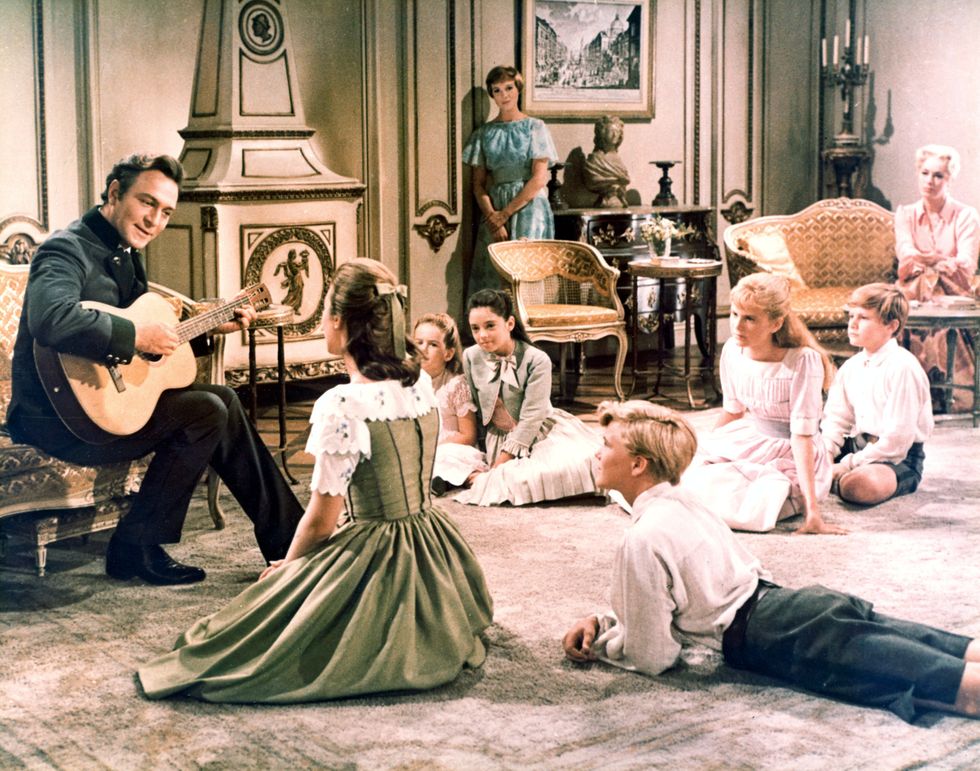 christopher plummer plays guitar and sings while seated in a living room, seven children sit or lie on the floor watching as julie andrews and another woman watch from across the room