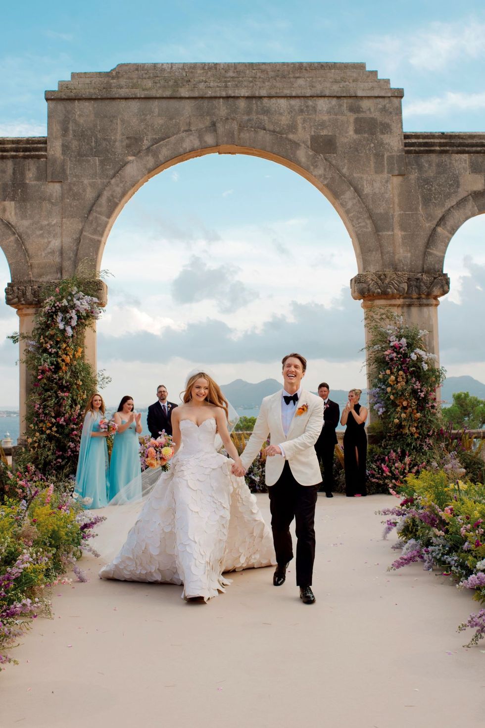a man and woman in wedding attire walking down a path with a large arch behind them