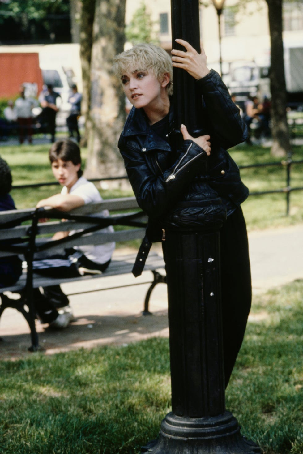 madonna holds on to a lamp post in a park with both hands as she stands behind it, looking towards the left of the camera frame