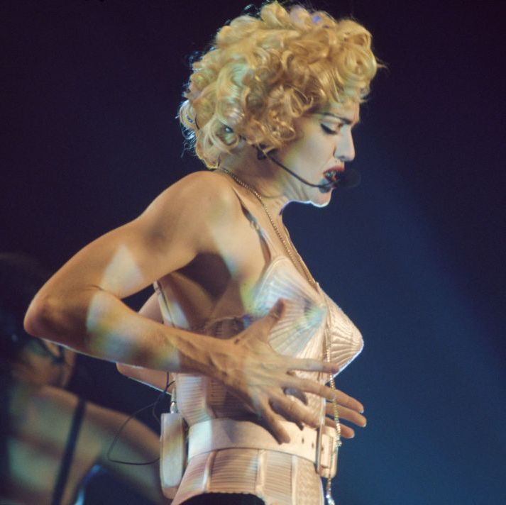 madonna, wearing a white outfit with her famous cone bra, singing into a headset microphone on a stage