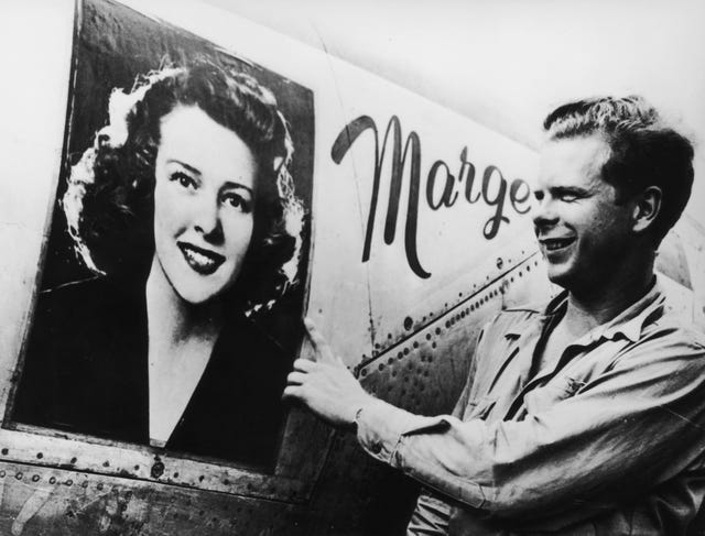 a man stands next to the side of a plane and smiles, he looks at a photo of a smiling woman on the plane, the word marge is written on the right