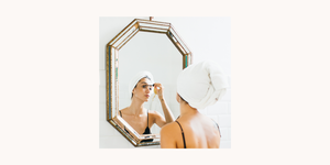 woman with a towel over her head applying mascara in the mirror