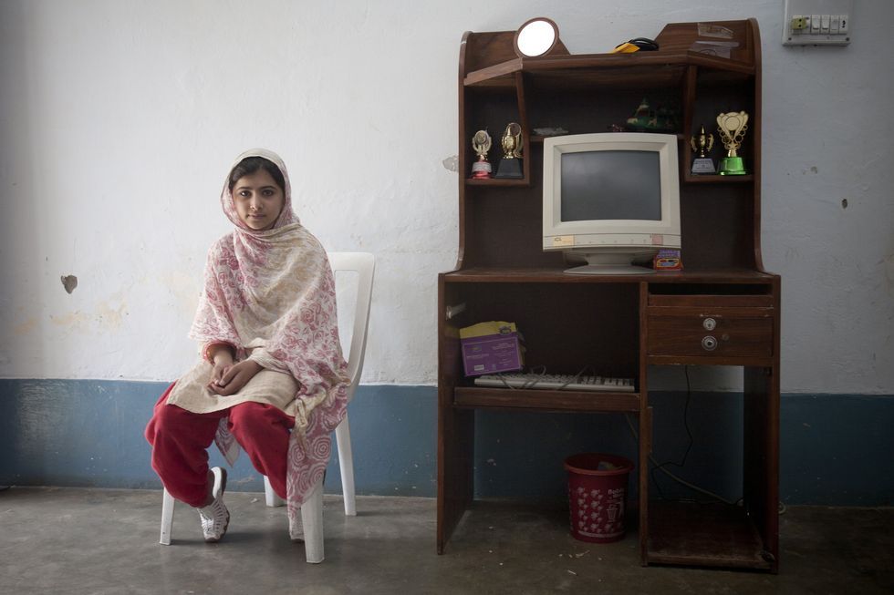 malala yousafzai sits on a white chair facing the camera, to the right is a wooden desk with a computer, medals, and other items