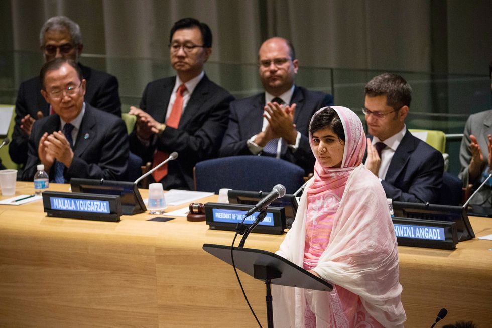 malala yousafzai stands at a podium with a microphone and looks outward as men sitting behind her clap, she wears a pink outfit