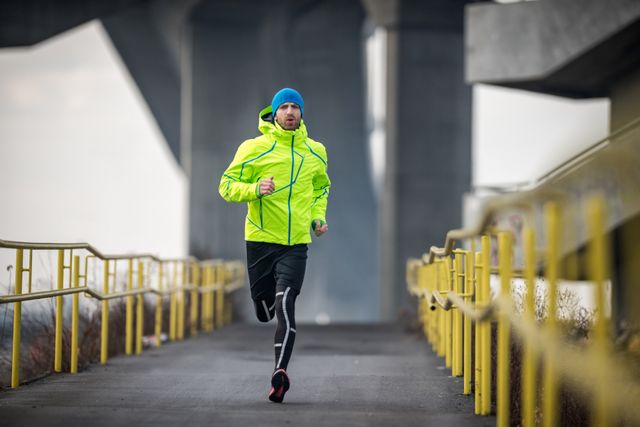 sportsman running over the bridge during his cardio routine on a winter day he is wearing green jacket and blue cap