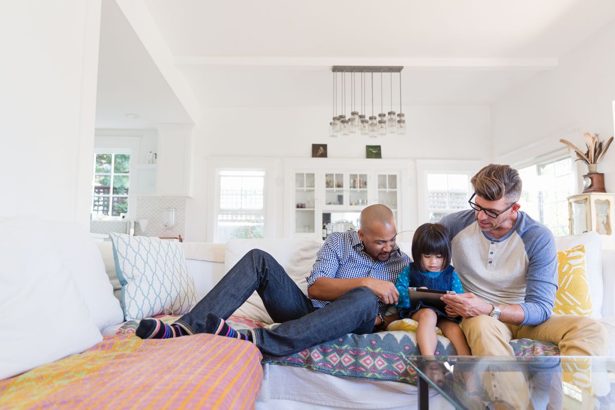 Male gay parents and adopted toddler daughter using digital tablet on living room sofa