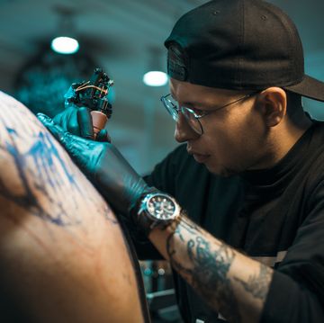 male tattoo master in black cap and glasses tattooing male back in tattoo salon