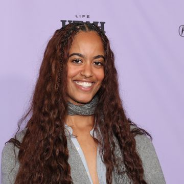 a woman smiles at the camera, she has long brown hair and wears a gray jacket and scarf