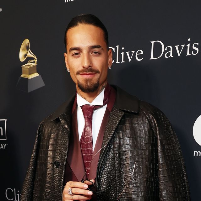 maluma looks at the camera while standing in front of a black background with logos, he wears a dark jacket over a maroon suit and tie with a white collared shirt, he wears a pair of glasses in his hand at his chest