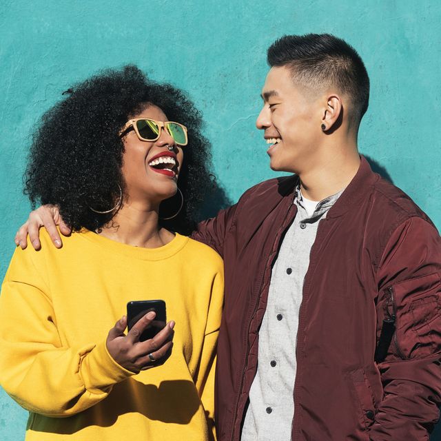 man and woman laughing while using mobile phone while standing against turquoise wall