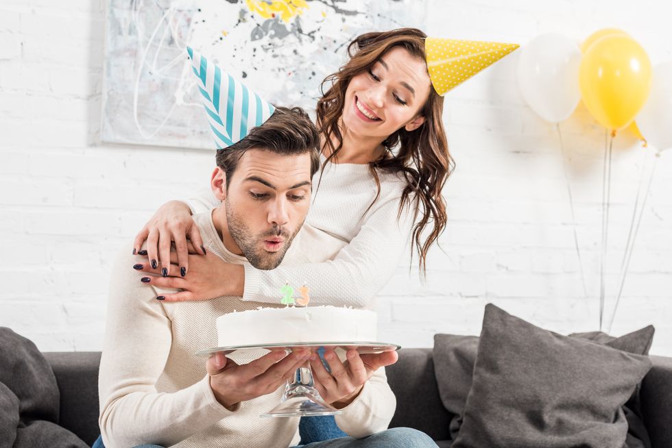 man blowing out candles on birthday cake with smiling woman in party hat on background