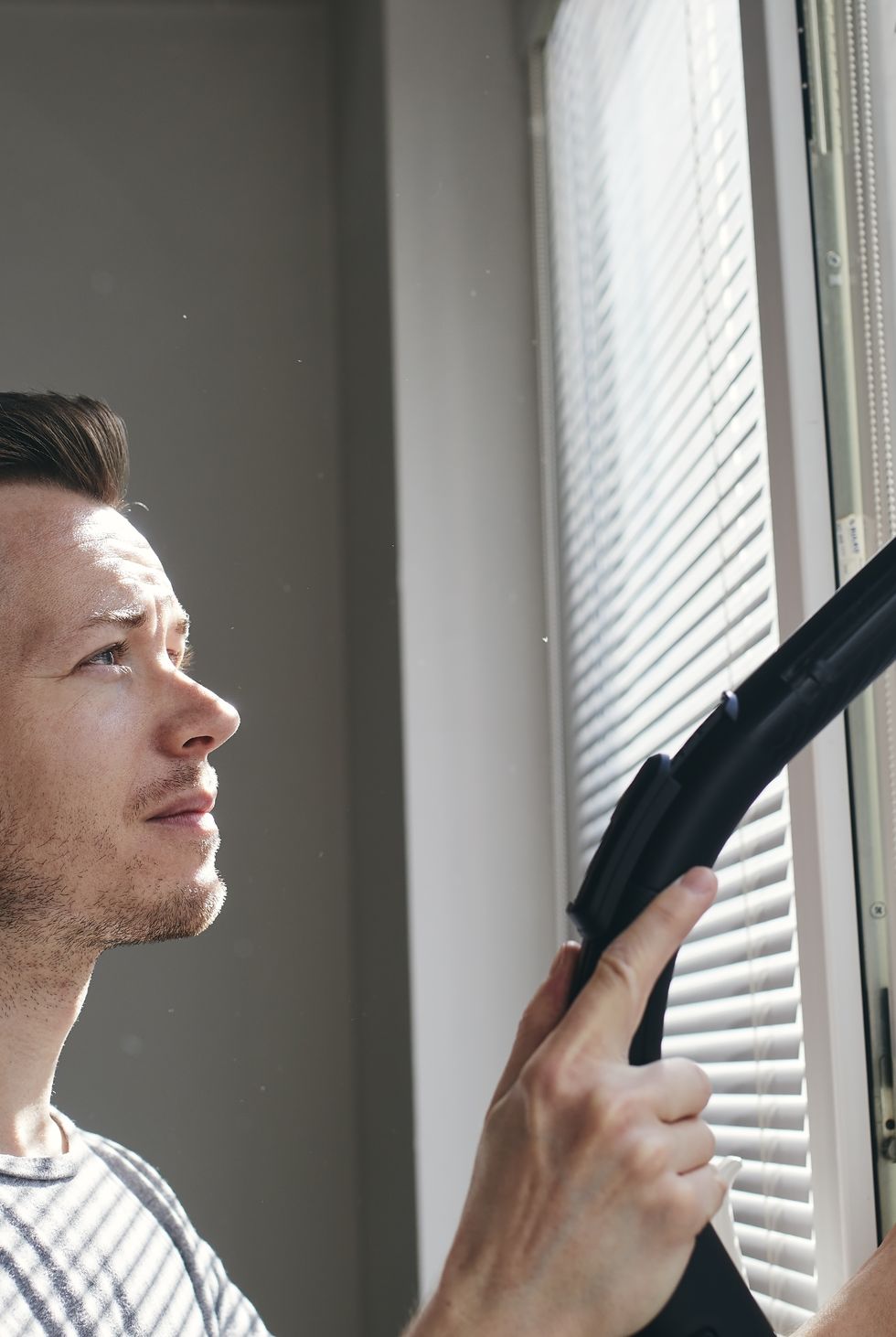 man cleaning dust from window blind by vacuum cleaner at home