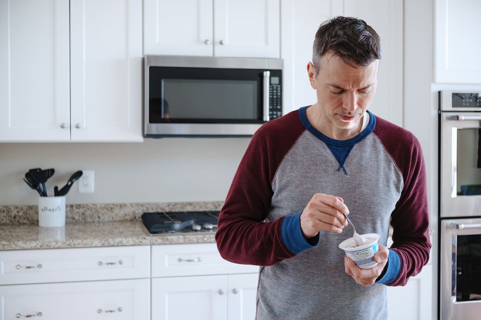 man eats a cup of yogurt while standing in kitchen
