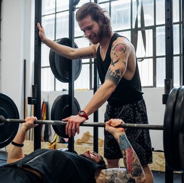 man lying on a weight bench about to bench press with another man standing with his hand on the bar