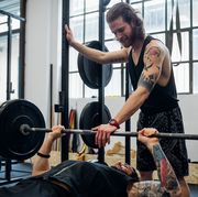 man lying on a weight bench about to bench press with another man standing with his hand on the bar