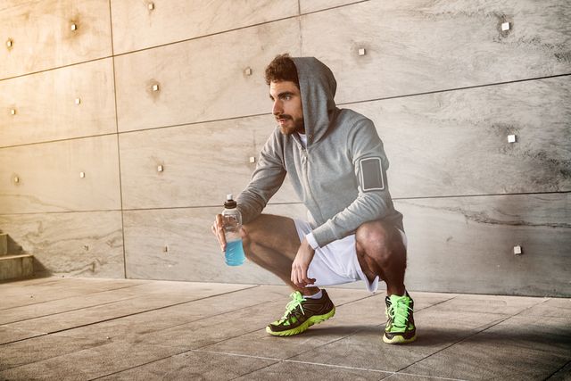 young man is resting crouched against a cocrete wall near a stadium entrance he's keeping an energy drink in his hand and catching his breath after intense workout