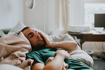 a man wakes up on a sofa, surrounded by sheets and duvets and pillows he rubs his eyes and face as he tries to rouse himself