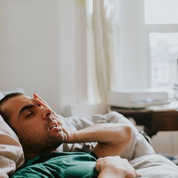 a man wakes up on a sofa, surrounded by sheets and duvets and pillows he rubs his eyes and face as he tries to rouse himself