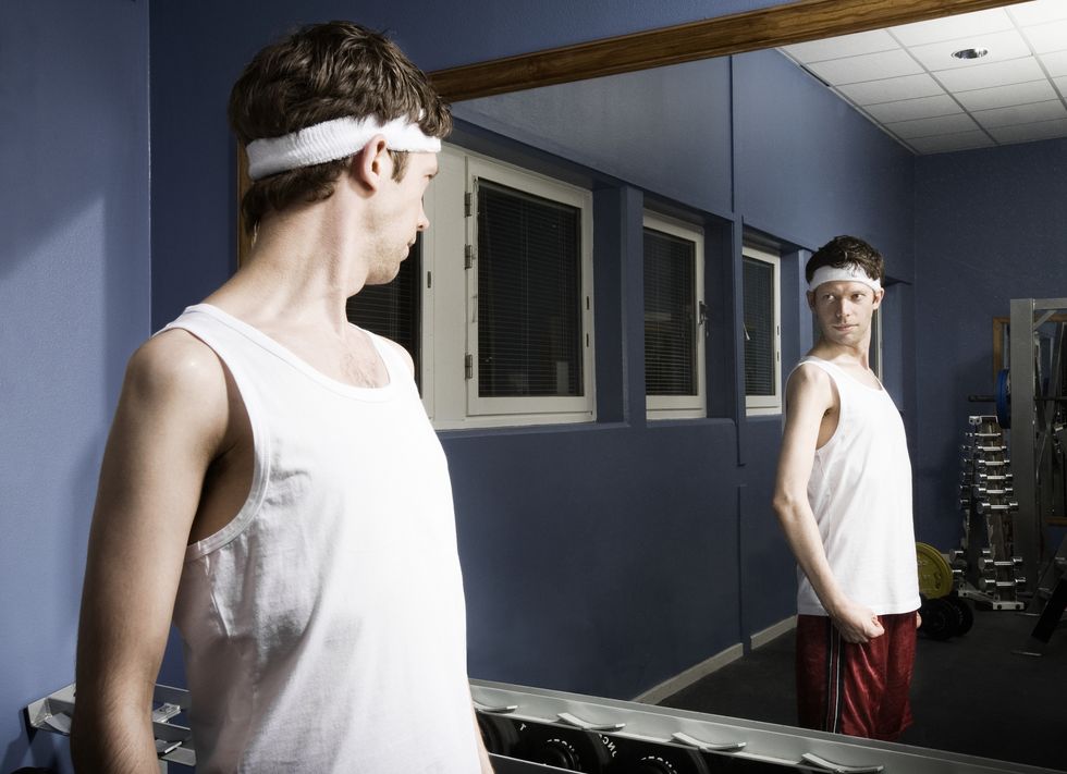 man wearing sweatband looking at reflection in gym mirror, flexing arm