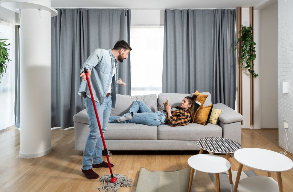 man wipes and cleans the floor of an apartment with mop and argues with a lazy woman lying on a sofa and spends time on social media on a smartphone end dont want to help him in housework