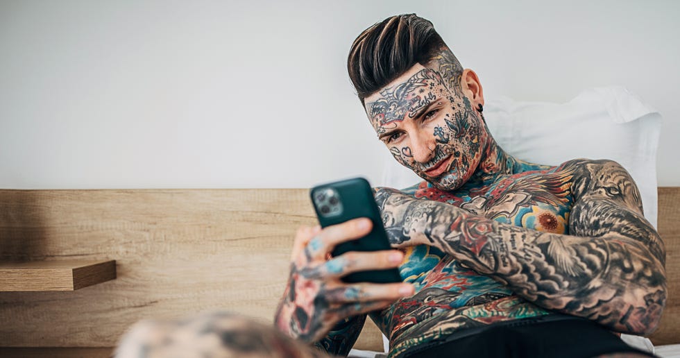 man with whole body covered in tattoos using smart phone in bedroom bed