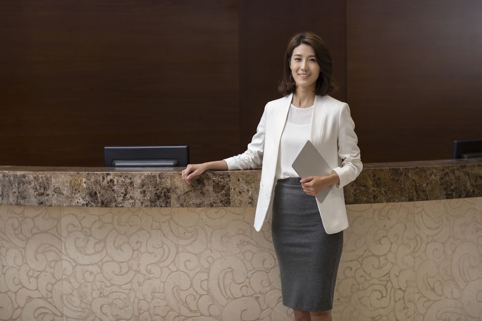la directora de un hotel posa en la recepción con el brazo apoyado en el mostrador y una tablet en la otra mano