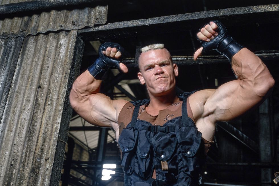 a man with huge arm muscles scowls at the camera as he flexes with his arms over his head, he wears tactical black gear and black gloves