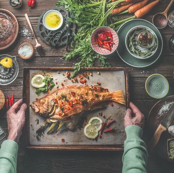man's hands holding roasted whole redfish stuffed with fennel and lemon on baking tray on rustic table with fresh ingredients and kitchen utensils