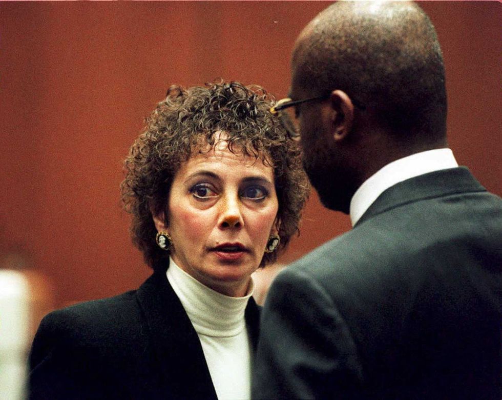 marcia clark looks past the camera as a man stands with his back to the camera, both wear dark suit jackets and collared shirts
