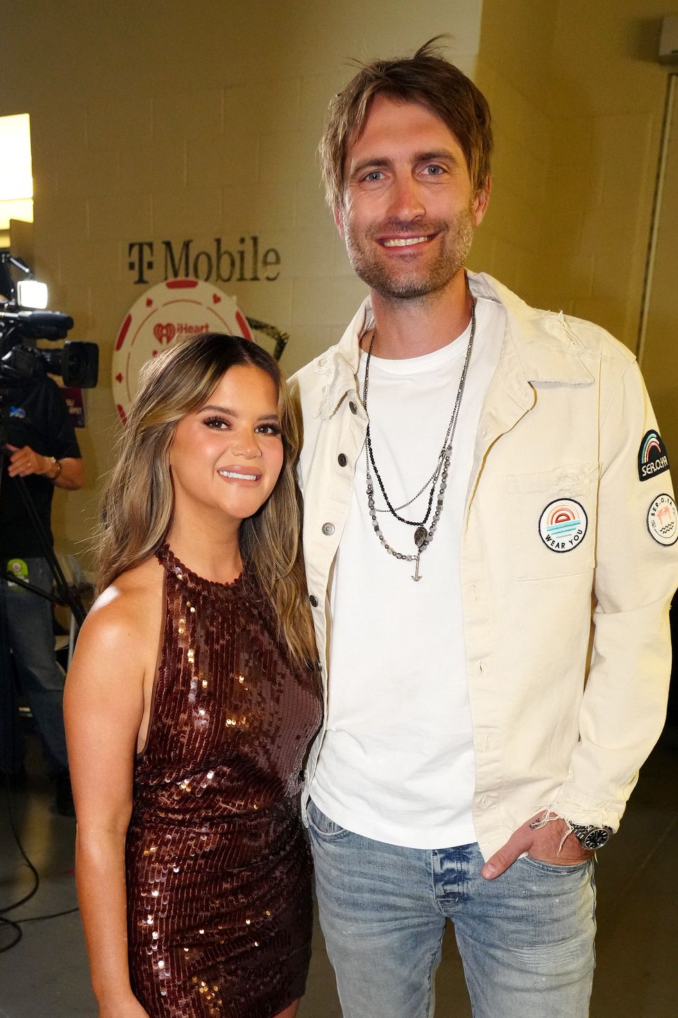 a woman and man stand inside embracing and smiling at the camera, she wears a brown sequin halter dress, he wears a white jacket white shirt and jeans