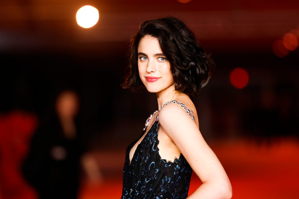 margaret qualley looks at the camera while standing in profile with one arm on her hip, she wears a sleeveless formal black gown with silver straps