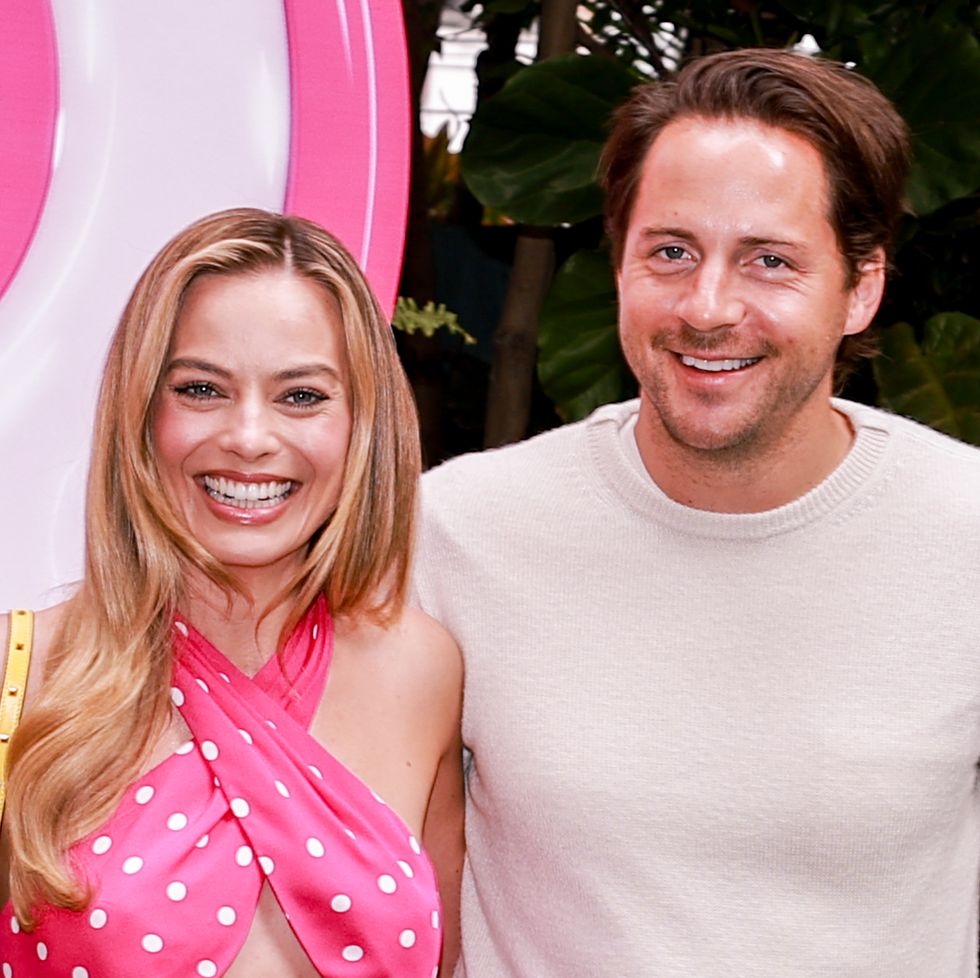 margot robbie and tom ackerley smiles at the camera while standing together, she wears a pink polka dotted top, he wears a cream colored sweater