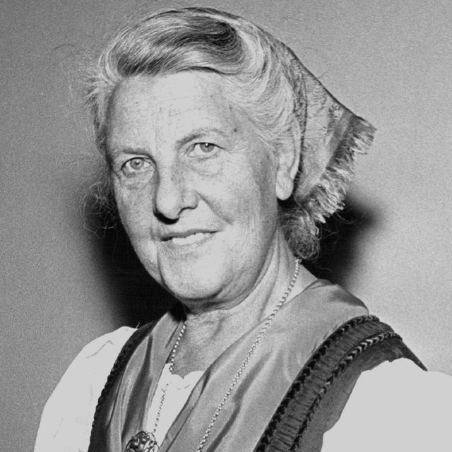 maria von trapp smiles at the camera, she wears a kerchief in her hair, a necklace, and a blouse