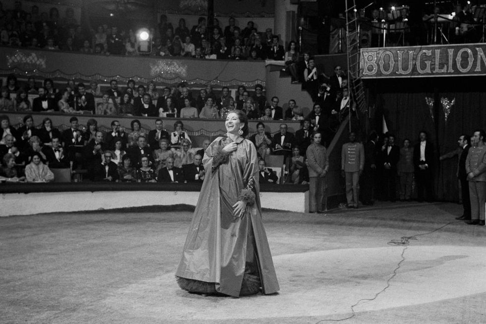 maria callas smiles while standing on a floor as audience members sit and watch, she wears a large robe style gown which she clutches around her