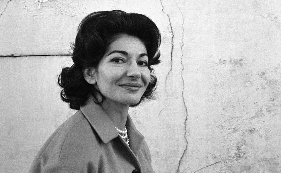 maria callas smiles at the camera, she walks to the right and wears a coat and pearl necklace