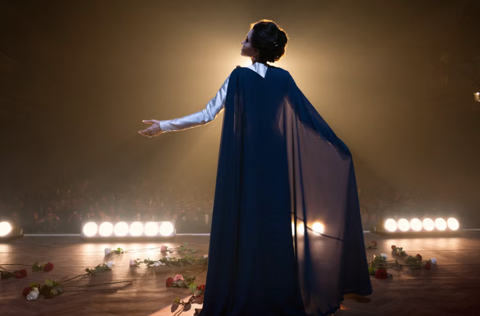 a performer dressed in a dark flowing cape and a white top extends their arms outward exuding an air of confidence and artistry